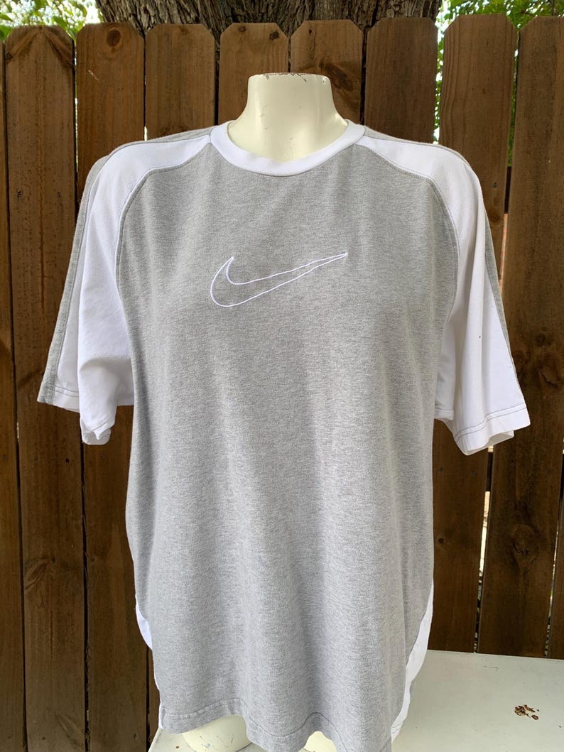 Puede incluir: Camiseta gris y blanca de manga corta con el logo de Nike bordado en el pecho. La camiseta tiene cuello redondo y mangas y paneles laterales blancos. El logo de Nike est&aacute; cosido en blanco.
