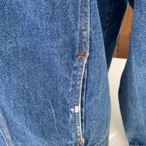 May include: Close-up of a blue denim jacket. The jacket features orange stitching and a pocket detail. The denim has a classic, worn look. The jacket's design is simple and functional.