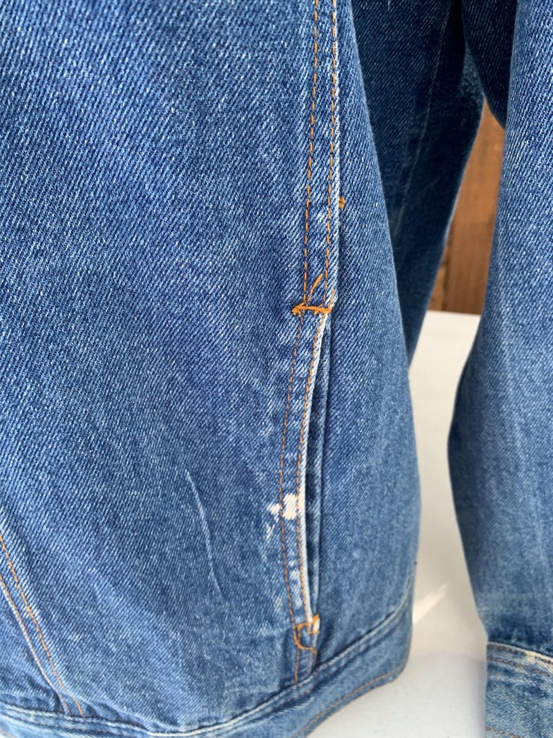 May include: Close-up of a blue denim jacket. The jacket features orange stitching and a pocket detail. The denim has a classic, worn look. The jacket's design is simple and functional.
