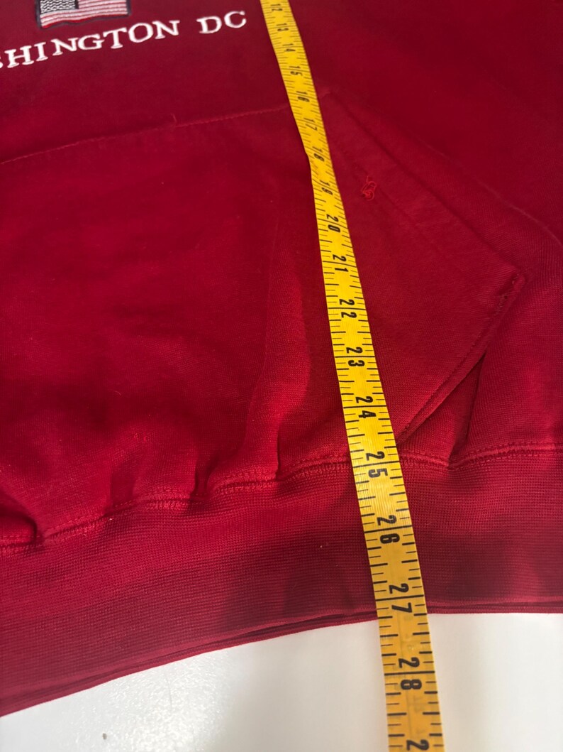 May include: A red hooded sweatshirt with the text "WASHINGTON DC" embroidered in white. A yellow measuring tape is placed over the sweatshirt, showing measurements in inches.