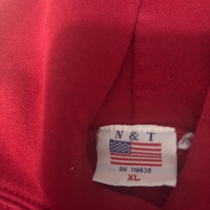 May include: A close-up of a red hoodie. The tag features the letters "N & T" above a small American flag, with "RN 118638" and "XL" printed below. The fabric appears to be a soft, knit material.