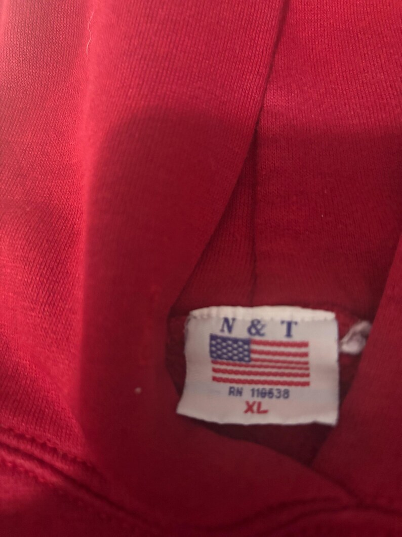 May include: A close-up of a red hoodie. The tag features the letters "N & T" above a small American flag, with "RN 118638" and "XL" printed below. The fabric appears to be a soft, knit material.