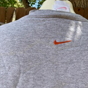 May include: A gray crew neck sweatshirt with an orange Nike swoosh logo embroidered on the chest. The sweatshirt has a ribbed collar and a tag on the back. The background is out of focus.