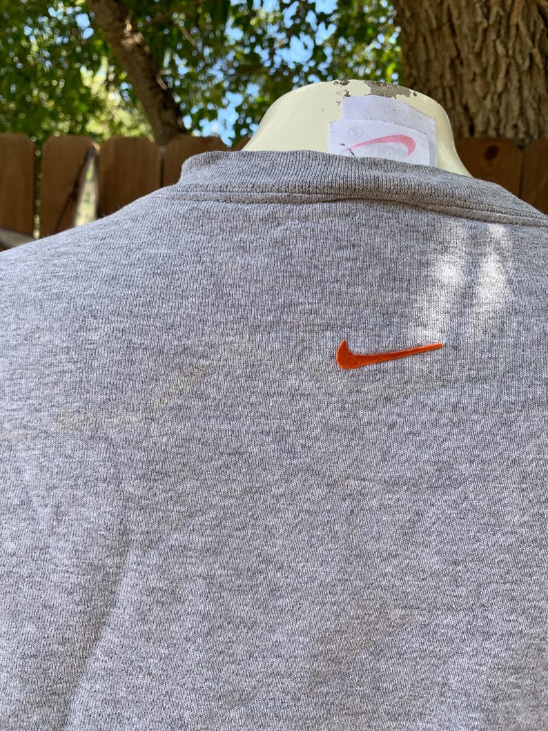 May include: A gray crew neck sweatshirt with an orange Nike swoosh logo embroidered on the chest. The sweatshirt has a ribbed collar and a tag on the back. The background is out of focus.