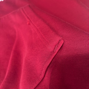 May include: Close-up of a red hoodie with a kangaroo pocket. The fabric is a solid red color with a visible texture. The stitching of the pocket is clearly visible. The top of the hoodie has white text that reads "ON DC".