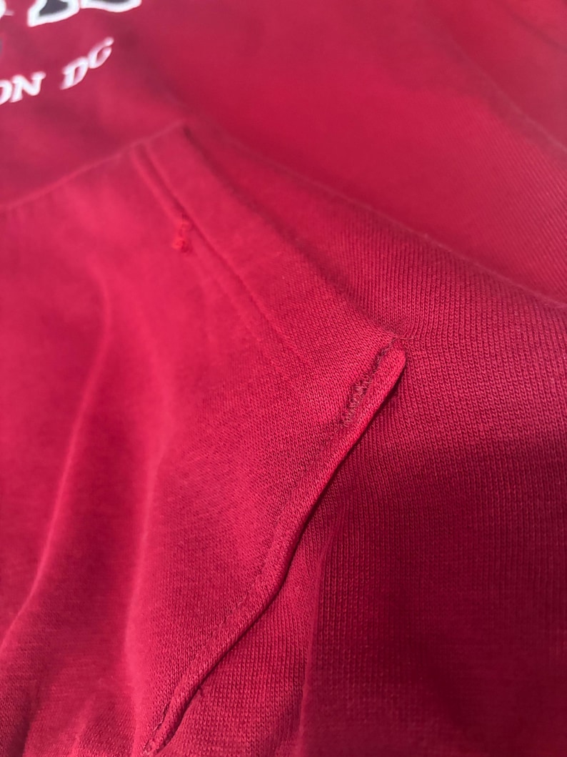 May include: Close-up of a red hoodie with a kangaroo pocket. The fabric is a solid red color with a visible texture. The stitching of the pocket is clearly visible. The top of the hoodie has white text that reads "ON DC".