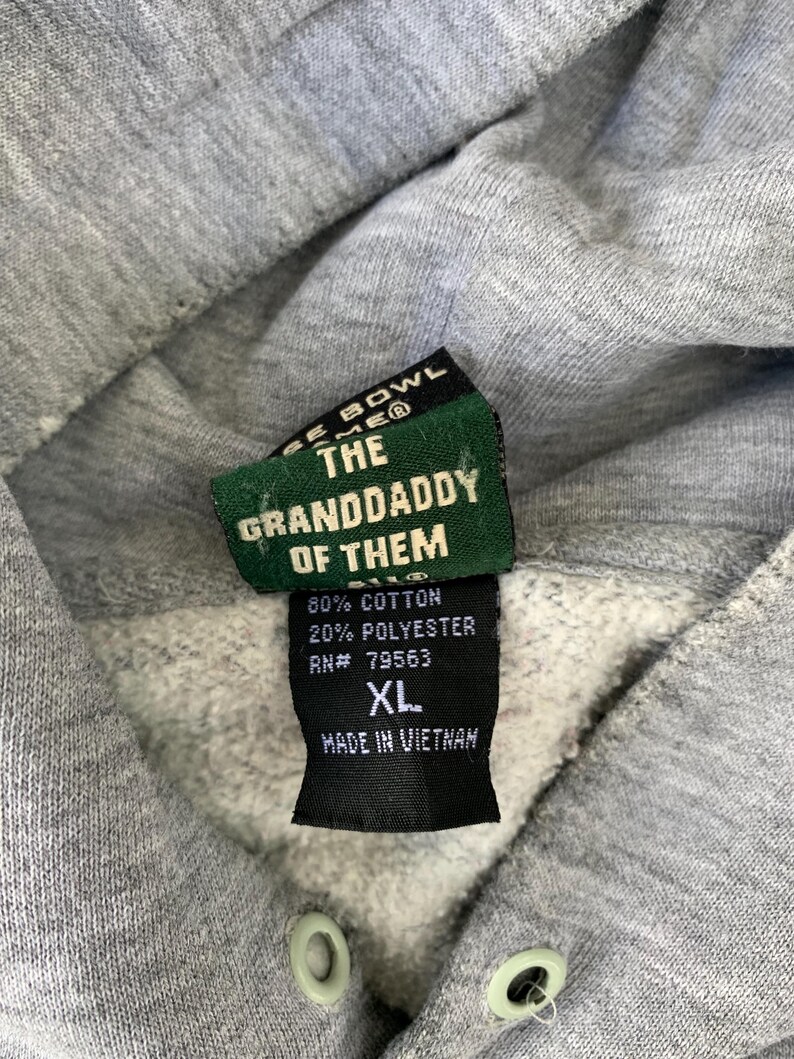 May include: Close-up of a gray hoodie with a tag that reads "THE GRANDDADDY OF THEM." The tag also indicates the hoodie is 80% cotton, 20% polyester, size XL, and made in Vietnam. The hoodie has a drawstring and a light gray color.