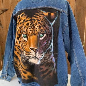 May include: A denim jacket with a large, detailed painting of a leopard's face on the back. The leopard has orange and black spots, green eyes, and a tan face. The jacket is a medium wash blue.