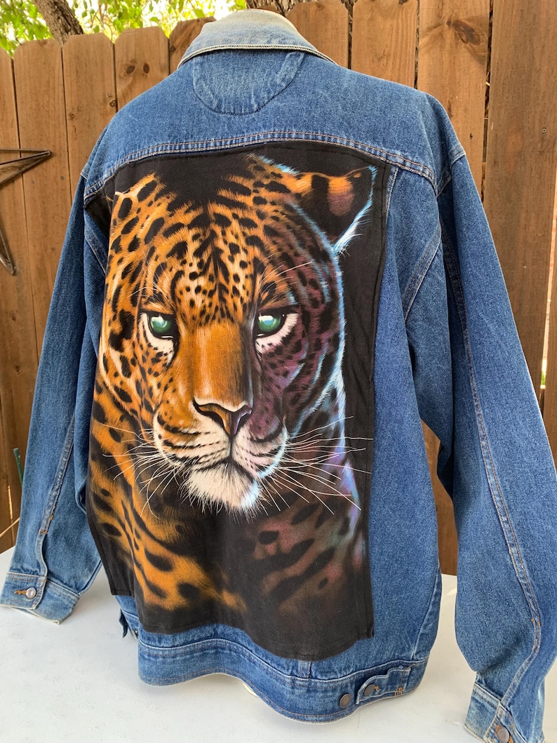 May include: A denim jacket with a large, detailed painting of a leopard's face on the back. The leopard has orange and black spots, green eyes, and a tan face. The jacket is a medium wash blue.