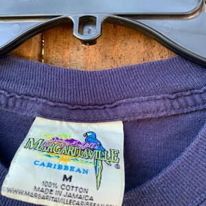 May include: A navy blue t-shirt with a white tag that says "Jimmy Buffett's Margaritaville Caribbean" and "100% Cotton Made in Jamaica" with a parrot graphic. The tag also says "M" for size.