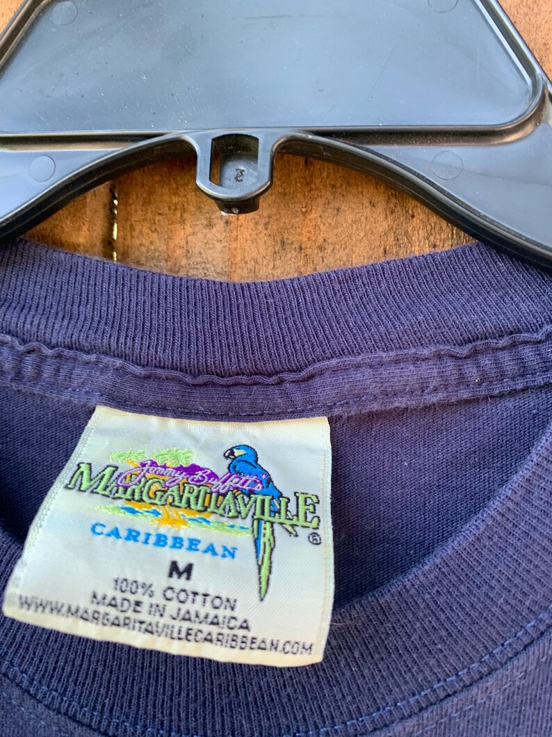 May include: A navy blue t-shirt with a white tag that says "Jimmy Buffett's Margaritaville Caribbean" and "100% Cotton Made in Jamaica" with a parrot graphic. The tag also says "M" for size.