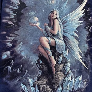 May include: A fantasy art print featuring a seated fairy with long silver hair, holding a glowing orb. The fairy has large, detailed blue wings and is surrounded by crystals and a starry night sky. The artwork is from the Anne Stokes Collection.