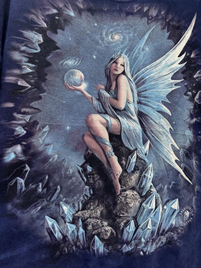 May include: A fantasy art print featuring a seated fairy with long silver hair, holding a glowing orb. The fairy has large, detailed blue wings and is surrounded by crystals and a starry night sky. The artwork is from the Anne Stokes Collection.