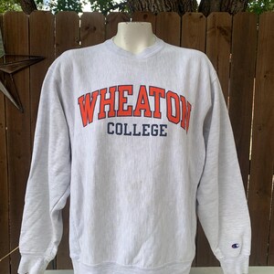 May include: A light gray sweatshirt with the text "WHEATON COLLEGE" in orange and navy blue. The sweatshirt has long sleeves and a Champion logo on the left sleeve cuff.