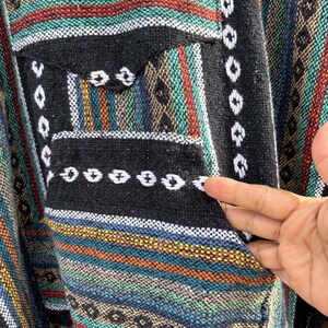 Pode incluir: Um poncho tecido colorido com um bolso preto. O poncho apresenta um padr&atilde;o listrado em tons de azul, vermelho, laranja e verde, com detalhes em branco e preto. O bolso &eacute; preto com costuras brancas.