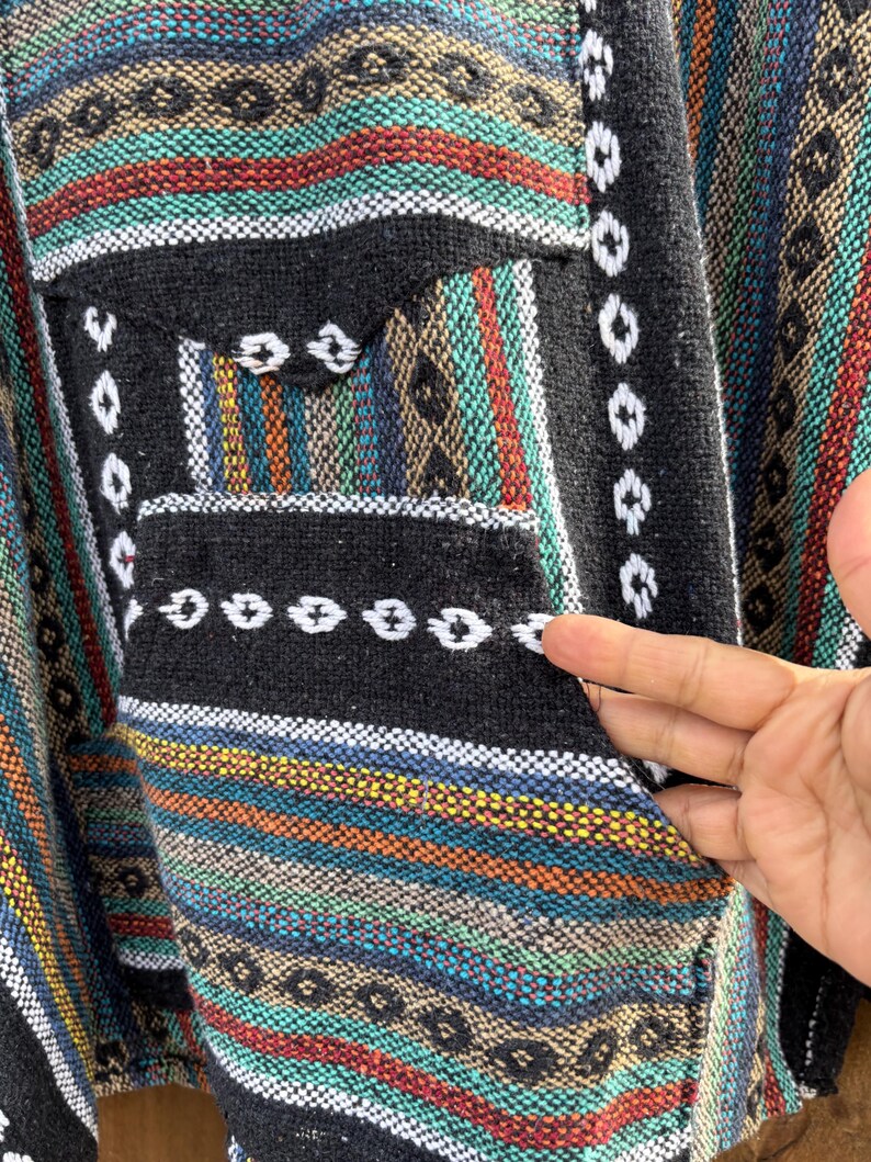 Pode incluir: Um poncho tecido colorido com um bolso preto. O poncho apresenta um padr&atilde;o listrado em tons de azul, vermelho, laranja e verde, com detalhes em branco e preto. O bolso &eacute; preto com costuras brancas.