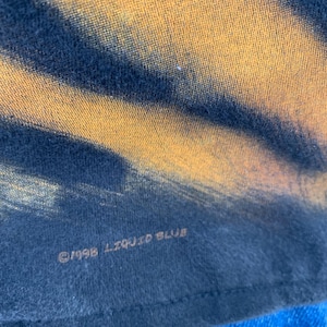 May include: Close-up of a tie-dye t-shirt with a yellow and black pattern. The text "&copy;1998 LIQUID BLUE" is printed on the bottom of the shirt. The shirt is made of a soft, comfortable material.