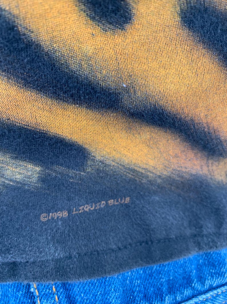 May include: Close-up of a tie-dye t-shirt with a yellow and black pattern. The text "&copy;1998 LIQUID BLUE" is printed on the bottom of the shirt. The shirt is made of a soft, comfortable material.