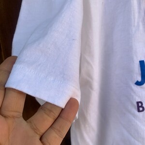 Pu&ograve; includere: T-shirt bianco con manica corta. La manica &egrave; a fuoco, mostrando l'orlo e la trama del tessuto. La maglietta ha testo blu e rosso sul davanti.