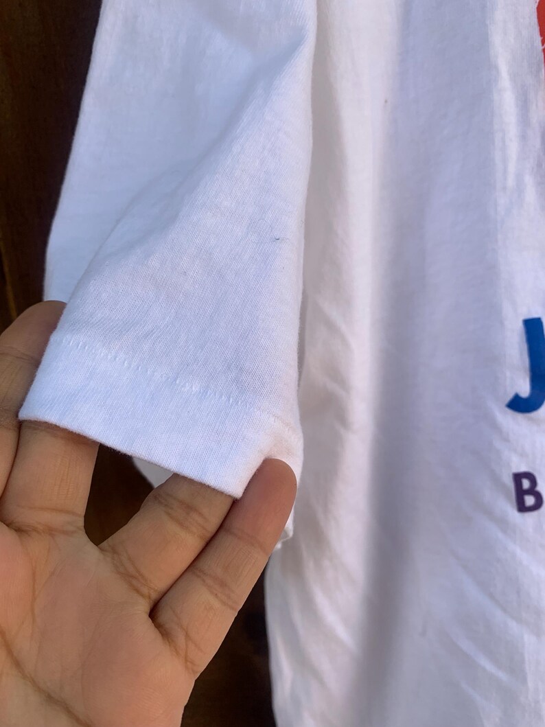 Pu&ograve; includere: T-shirt bianco con manica corta. La manica &egrave; a fuoco, mostrando l'orlo e la trama del tessuto. La maglietta ha testo blu e rosso sul davanti.