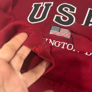 May include: A red sweatshirt with the word "USA" in white letters with a black outline. Below the letters is a small American flag. The word "Washington" is also visible. The sleeve has a tear.