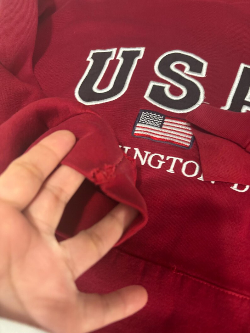 May include: A red sweatshirt with the word "USA" in white letters with a black outline. Below the letters is a small American flag. The word "Washington" is also visible. The sleeve has a tear.