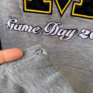 May include: Gray sweatshirt with the text "Game Day 20" and a large "M" logo in navy blue and yellow. The sweatshirt has a distressed appearance with a tear near the shoulder.