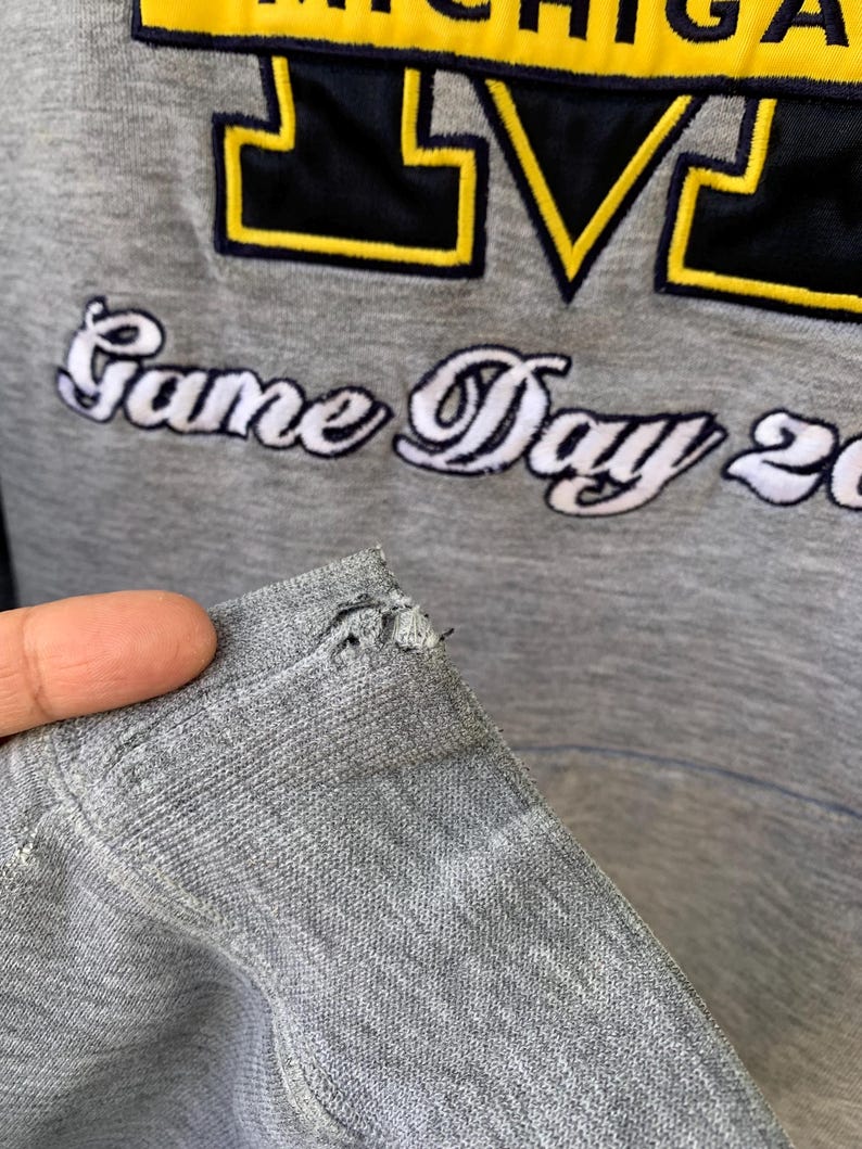May include: Gray sweatshirt with the text "Game Day 20" and a large "M" logo in navy blue and yellow. The sweatshirt has a distressed appearance with a tear near the shoulder.