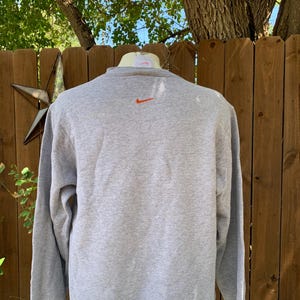 May include: A gray sweatshirt with an orange Nike logo on the upper back. The long-sleeved crew neck shirt is made of a soft material. The background includes a wooden fence, a star-shaped decoration, and tree branches.
