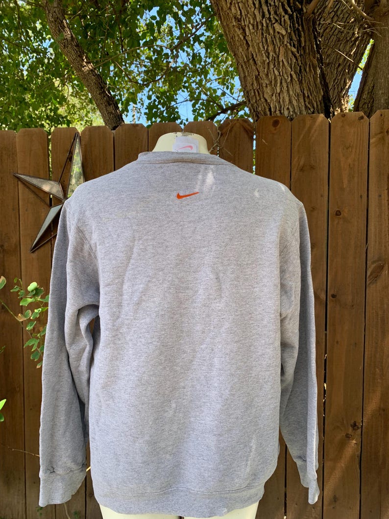 May include: A gray sweatshirt with an orange Nike logo on the upper back. The long-sleeved crew neck shirt is made of a soft material. The background includes a wooden fence, a star-shaped decoration, and tree branches.