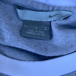 Puede incluir: Primer plano de una camiseta gris con cuello blanco. La etiqueta dice "MADE IN INDIA FABRIQUE EN INDE" con el logo de Nike. La camiseta es talla L.