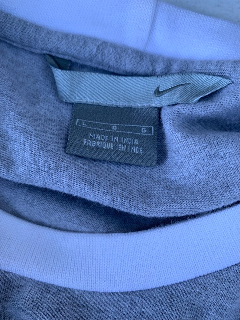 Puede incluir: Primer plano de una camiseta gris con cuello blanco. La etiqueta dice "MADE IN INDIA FABRIQUE EN INDE" con el logo de Nike. La camiseta es talla L.