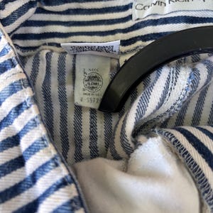 Puede incluir: Primer plano de una prenda a rayas azules y blancas. La etiqueta Calvin Klein Sport es visible. La prenda tiene una etiqueta con texto y un logotipo. La tela es de un material de mezclilla clara.