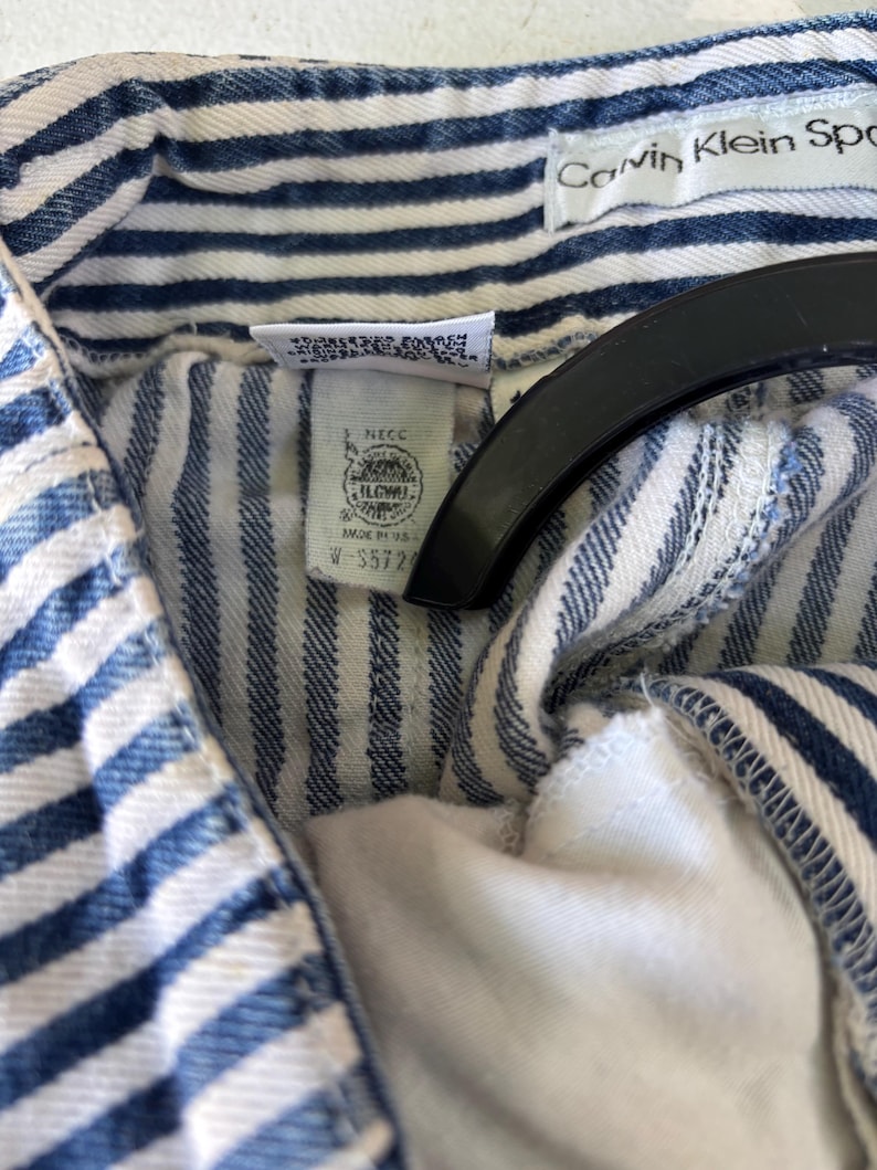 Puede incluir: Primer plano de una prenda a rayas azules y blancas. La etiqueta Calvin Klein Sport es visible. La prenda tiene una etiqueta con texto y un logotipo. La tela es de un material de mezclilla clara.