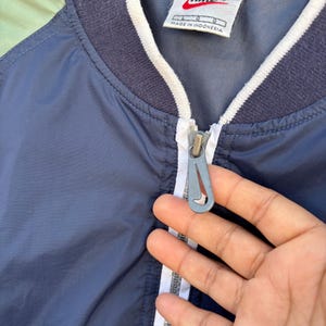 May include: A navy blue Nike jacket with a white zipper and trim. The jacket has a white collar and a tag that says "Nike" and "Made in Indonesia". The jacket is a size L.