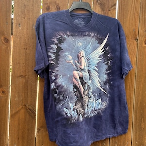 May include: Dark blue t-shirt featuring a fantasy art print. The graphic depicts a fairy with blue wings, holding a glowing orb, surrounded by crystals and a starry background. The Mountain brand logo is visible.
