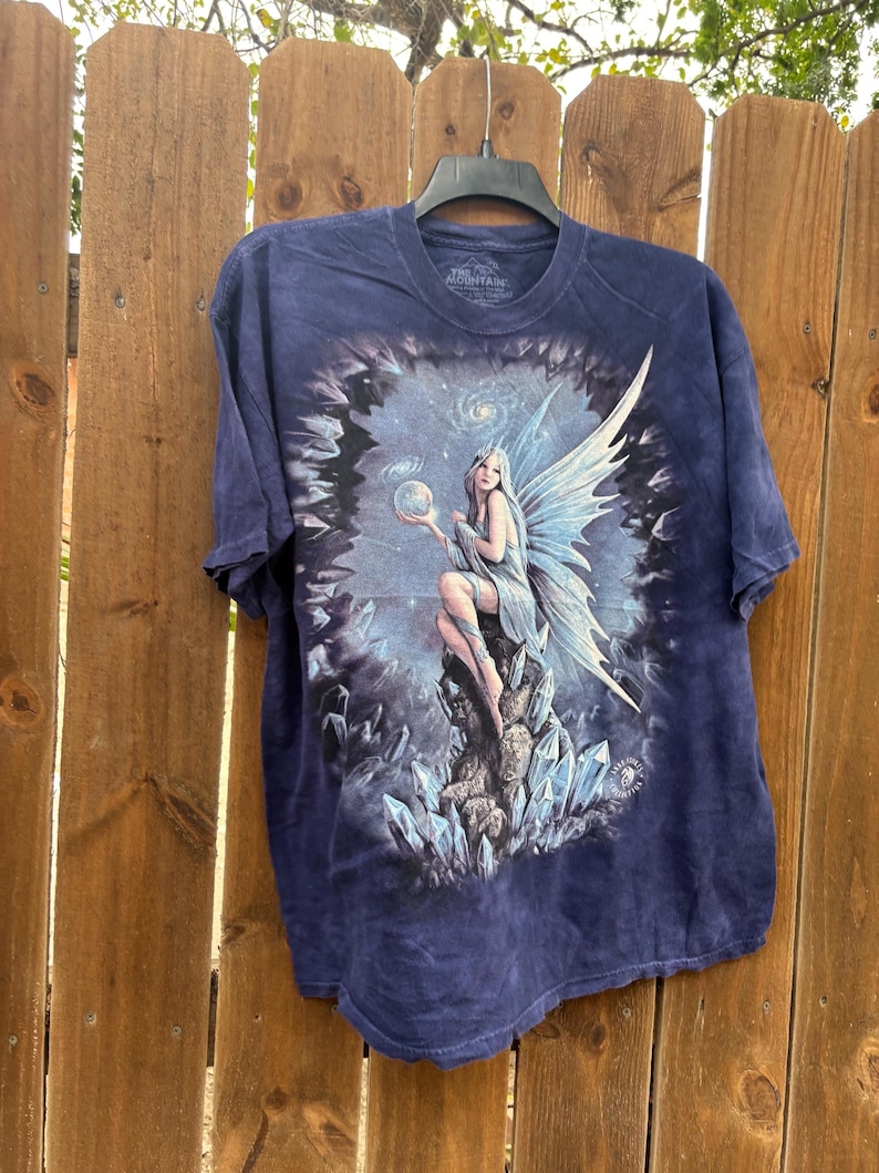 May include: Dark blue t-shirt featuring a fantasy art print. The graphic depicts a fairy with blue wings, holding a glowing orb, surrounded by crystals and a starry background. The Mountain brand logo is visible.