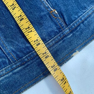May include: Close-up of a blue denim jacket with visible stitching and a yellow measuring tape. The tape shows measurements from 24 to 33 inches. The jacket has a classic design.