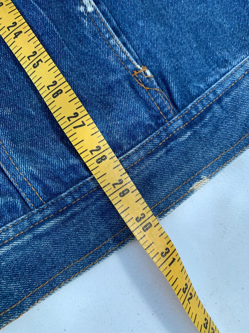 May include: Close-up of a blue denim jacket with visible stitching and a yellow measuring tape. The tape shows measurements from 24 to 33 inches. The jacket has a classic design.