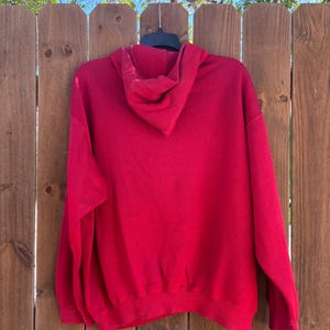 May include: A solid red hoodie with a hood and long sleeves. The hoodie is hanging on a black hanger against a wooden fence. The hoodie is made of a soft material.