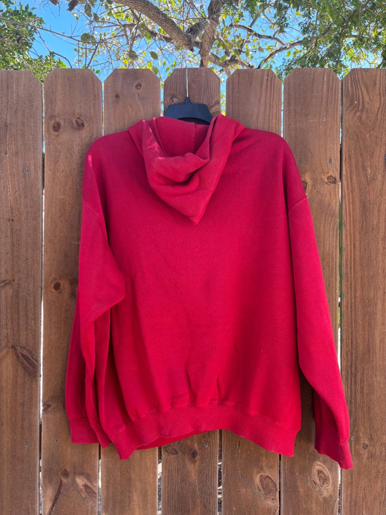 May include: A solid red hoodie with a hood and long sleeves. The hoodie is hanging on a black hanger against a wooden fence. The hoodie is made of a soft material.