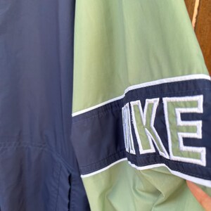 May include: A navy blue and green Nike jacket. The jacket features a color block design with the Nike logo in white and green on the sleeve. The jacket has a white stripe detail.