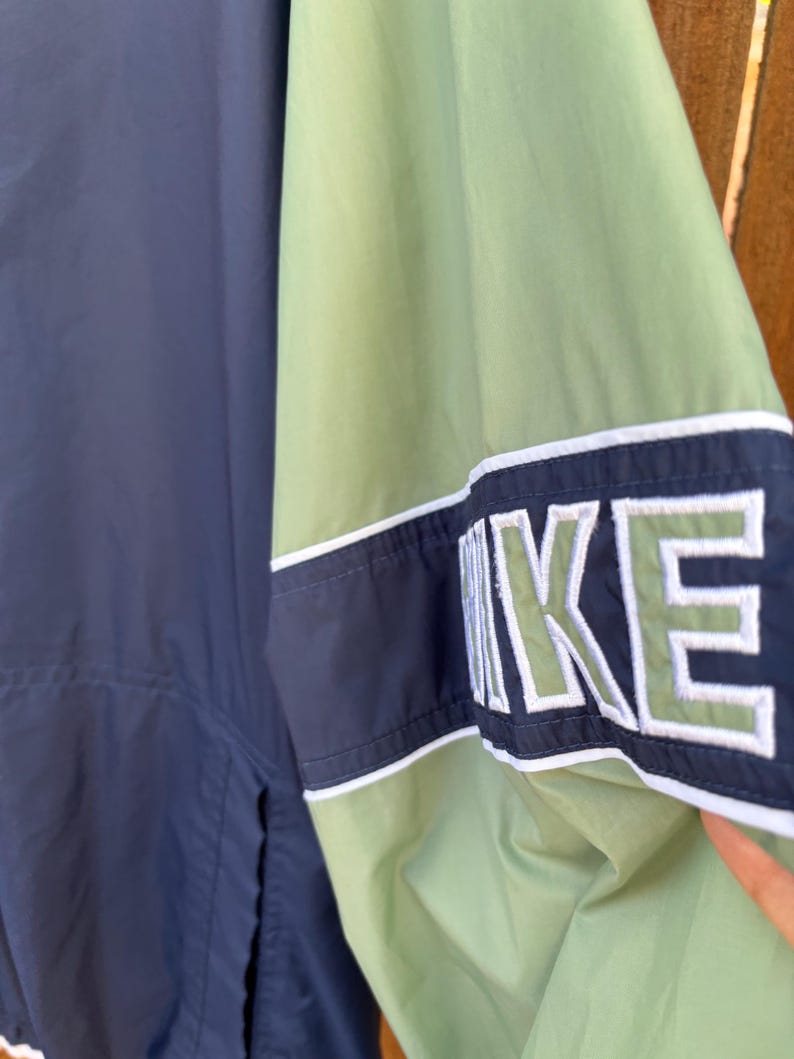 May include: A navy blue and green Nike jacket. The jacket features a color block design with the Nike logo in white and green on the sleeve. The jacket has a white stripe detail.