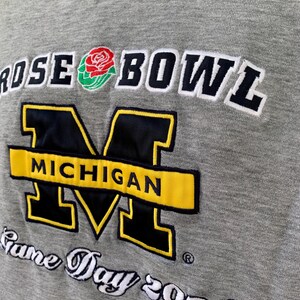 May include: Gray sweatshirt with "ROSE BOWL" and "MICHIGAN" embroidered in black, yellow, and white. Features a rose graphic and "Game Day 2004" text. A vintage sports apparel item.
