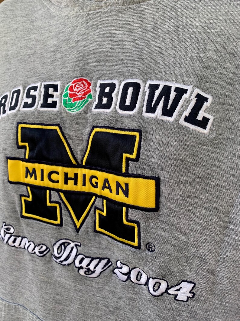May include: Gray sweatshirt with "ROSE BOWL" and "MICHIGAN" embroidered in black, yellow, and white. Features a rose graphic and "Game Day 2004" text. A vintage sports apparel item.