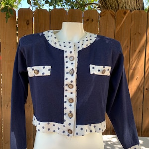 May include: A navy blue cropped cardigan with white polka dots. The cardigan has a button closure and two pockets. The sleeves are long and have a white polka dot trim.