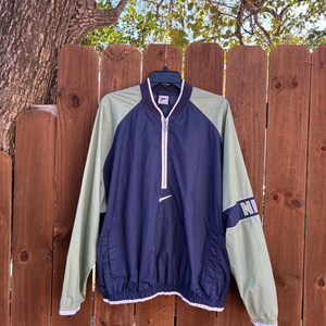 May include: A vintage Nike windbreaker jacket in navy blue and light green. The jacket features a white zipper, a small Nike logo, and the word "NI" on the sleeve. The jacket has a drawstring hem and white trim.