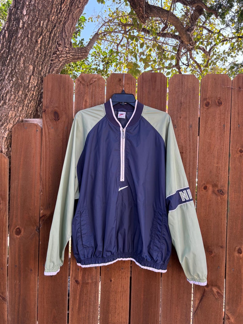 May include: A vintage Nike windbreaker jacket in navy blue and light green. The jacket features a white zipper, a small Nike logo, and the word "NI" on the sleeve. The jacket has a drawstring hem and white trim.