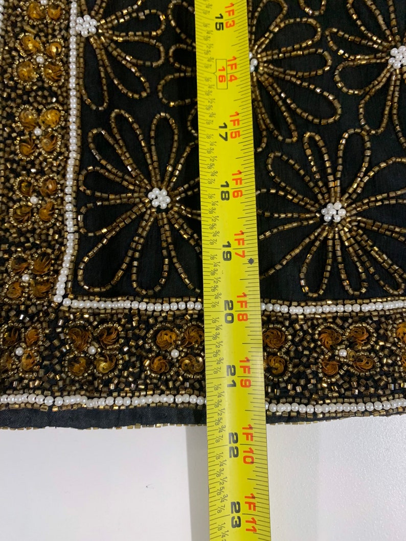 May include: Black fabric with gold and white beading in a floral pattern. A yellow measuring tape is to the right of the fabric, showing a measurement of 23 inches.