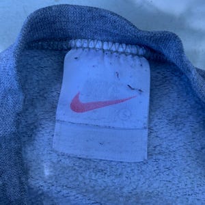 May include: Close-up of a gray sweatshirt collar with a white tag. The tag features a pink Nike swoosh logo. The fabric has a textured, fleecy appearance.